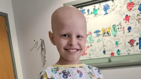 A smiling boy in a hospital room wearing a white and yellow children's hospital gown standing in front of a poster of cartoon characters.