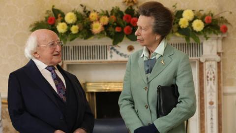 A small man with white hair and glasses in a navy suit standing beside a slim woman in Cambridge blue blazer in a royal looking palace with a fireplace and Irish flag behind them. 