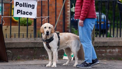 A white dog on a lead is held by owner wearing a red jacket and blur jeans outside a sign saying 'polling place' in Burrelton, Scotland on 7 May