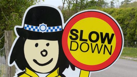 A sign outside a school with a cartoon-like depiction of a lollipop lady with the words "slow down" written on the lollipop.