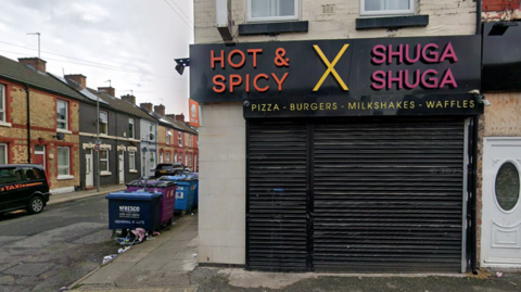 A shop has a sign saying 'Hot and Spicy', 'X' and 'Shuga Shuga' with 'pizzas - burgers - milkshake - waffles' underneath. Black shutters are down across the shop front. Industrial waste bins can be seen in a side street.