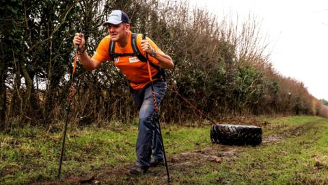 Jordan Wylie, carrying two hiking poles, drags a large tyre across a field. He wears grey trousers, an orange t-shirt and a grey cap.