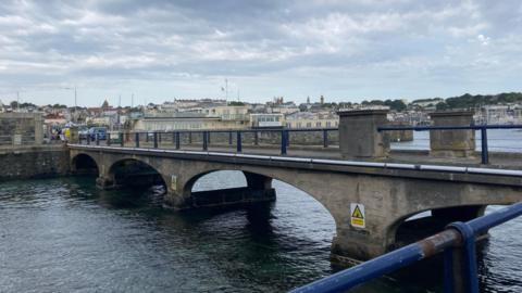 The Castle Cornet Bridge in Guernsey. It is stone and has blue railings. There are some danger signs on the side of it. There is also the sea underneath it. 