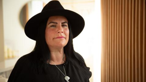 A woman wearing a black hat and a black top, standing near a window and looking at the camera