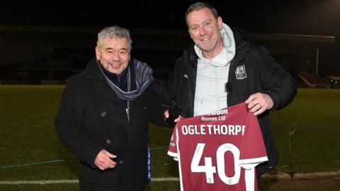 A man in a navy pea coat and a blue and grey scarf stands next to a man in a grey hoodie and black puffer jacket holding a claret football shirt with "Oglethorpe 40" in white letters.