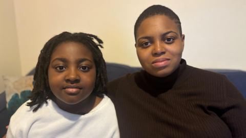 A young girl with dark hair in plaits and a woman with short dark hair in a dark top sitting on a sofa in front of a pale wall.