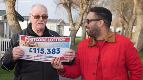 A man in a red coat passing a Postcode Lottery cheque to an older man in a black coat. The cheque says £115,383 on it.