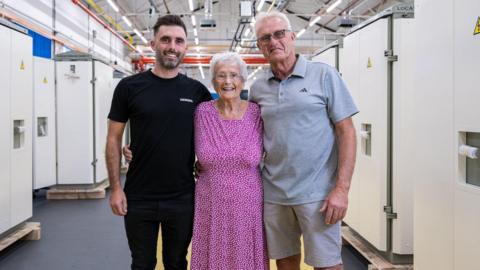 A smiling grandma with short white hair and wearing a fuschia polka dot dress, standing arm in arm with a middle-aged man to the right and a younger man with stubble to the left. They all look happy and are in an industrial-looking warehouse.