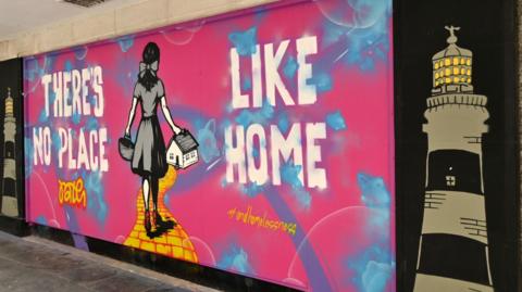 A painted mural. The two sides are painted back with a black and grey lighthouse and yellow light coming out of them. The main mural is pink with blue streaks and bubbles. There is white writing that says "There's no place like home" and yellow writing which says "PATH" and  "#endhomelessness". A painting of Dorothy, facing away, holding a basket in her left hand and house in her right hand can be seen walking down the yellow brick road. 