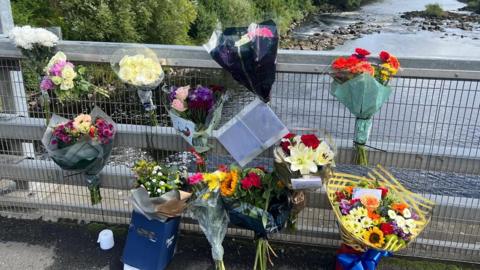 Eleven bouquets of flowers are tied to the metal railings of a bridge over a river. They are of various bright colours and wrapped in plastic. There is also a white card attached, but the writing is too small to be made out. There are tall, green trees bordering the water below.