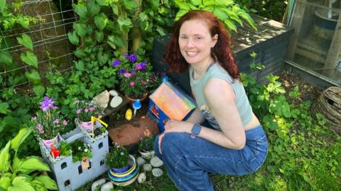 Christie Barnes, a woman with red hair wearing blue jeans, squats down next to a miniature castle and house for mice in her back garden. 