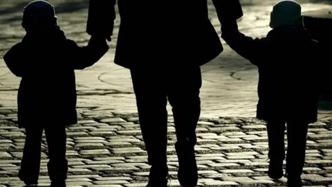 An adult walks on a cobbled street with a child either side of them. They are each holding hands in the silhouetted image.