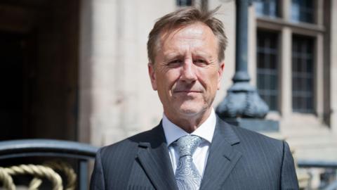 A man with brown hair, a grey suit and pale grey tie with standing in front of a sandstone building.