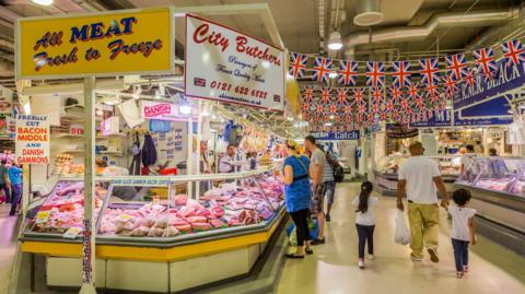 An indoor market, with traders and shoppers at a butcher's counter on the left and a man and two children walking past. Flags can be seen hanging from the ceiling and signs advertising bacon, gammon and other meats. other stalls are visible on the right.