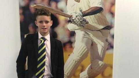 A schoolboy in a black blazer, white shirt and navy and yellow diagonally striped tie standing in front of a poster showing a cricketer.