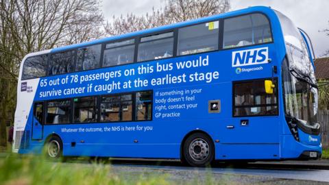 A blue and white double decker bus with the NHS logo emblazoned on the top deck. The words  "65 out of 78 passengers on this bus would survive cancer if caught at the earliest stage" are also painted on the top deck

Below, a caption reads: "if something in your body doesn't feel right, contact your GP practice, whatever the outcome, the NHS is here for you."