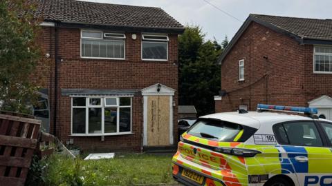The exterior of a house with a boarded up door. There is a police car parked outside.