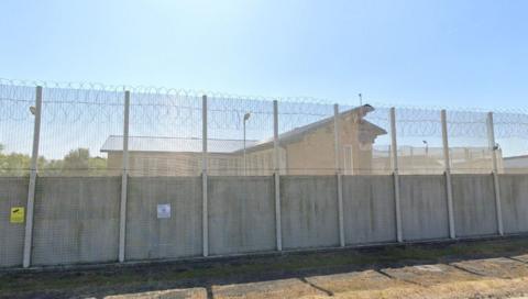 Streetview of a large prison fence with buildings beyond