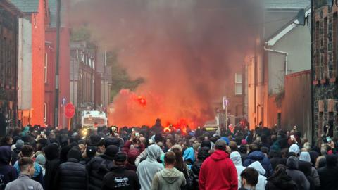 People on the streets during Ballymena protest. A large crowd. There are buildings along the side of the street and a police van in the distance. There is a white police van. Smoke is rising into the air.