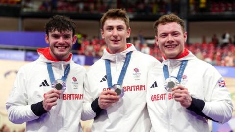 Ed Lowe, Hamish Turnbull and Jack Carlin all with short hair, smiling into the camera and holding their silver medals in white Great Britain hoodies