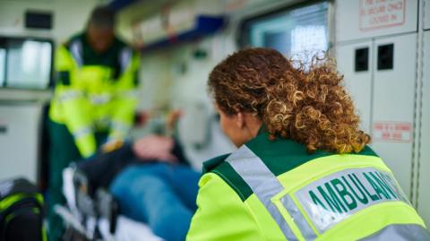 Two paramedics load a stretcher bearing a patient into an ambulance - one, in the foreground, is wearing a yellow luminous jacket with the word Ambulance on the back of it.