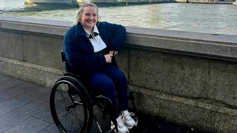 Chloe Docker sitting in her wheelchair pictured by the River Thames in London. She has blonde hair and is wearing a white top and a dark-blue cardigan and jeans.
