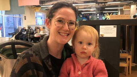 Judith Crozier and her two-year-old daughter in the children's play area of  2 Royal Avenue.   Judith has dark hair tied back and glasses.  She is wearing a dark, patterned jacket and a black top. Her daughter has blonde hair and is wearing a pink cardigan. 