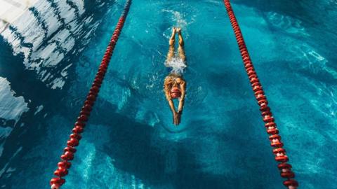 An image of a swimming pool from above - showing a female swimmer in a central lane mid-stroke, her arms outstretched in front of her. Red lane dividers are either side of her.