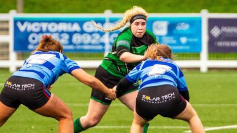 A blonde woman in a green top playing rugby. She had the bball and is about to try passing two opposing players in light and dark blue hooped tops.