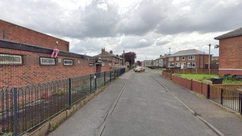 Millbrook Avenue is a residential road. A red-bricked social club is on the left, with a blue metal fence in front of it. Two-storey red bricked semi-detached houses line the road further down.