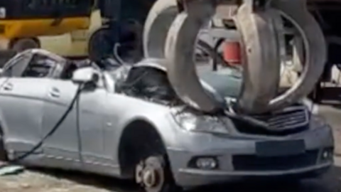 Silver car being crushed with large grabber.