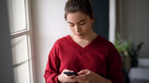 A disappointed-looking young woman in a red v-neck jumper looking at her phone.