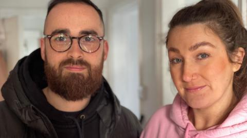 Head and shoulders shot of Emma Woolley and her husband, Jakob. She has dark hair and is wearing a pink hoodie. He has glasses and a beard and is wearing a dark-coloured jacket.