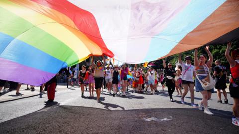 A crowd of people holding a giant pride flag above their heads. The flag stretches the length of the photograph. They are walking along a street and there are more people in the background.