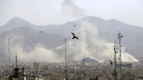 plumes of smoke rise over Tehran in Iran, with mountains seen silhouetted in the background.