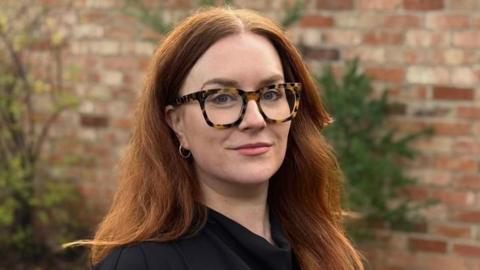 Matilda Robinson-Murphy has long auburn hair. She is standing in front of a brick wall, wearing a black top and glasses with tortoise shell rims.
