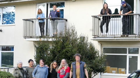 Artists standing outside Commercial Road Studios. Six of them are on the road in front of the house while four of them are waving from the building's balconies.