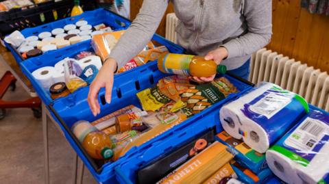 A line of blue plastic boxes with food and toilet roll in them. A person in a grey top is holding a bottle of squash and has another hand hovered over the boxes.
