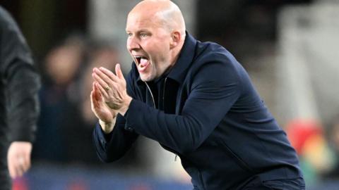 Henrik Pedersen crouches on touchline, hands clapping encouragement as he shouts instructions, wearing all navy blue trousers and zipped warm jacket.