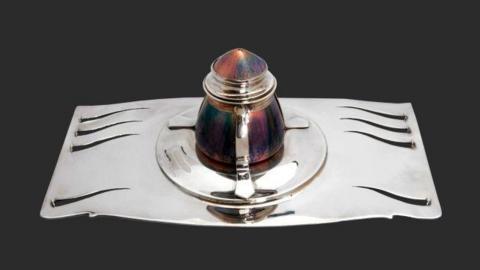 The inkwell, which has a decorative silver base with a colourful pot that has a lid in the middle of it.