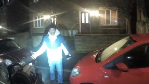 A man lit up by white light standing outside a house with his arms outstretched.