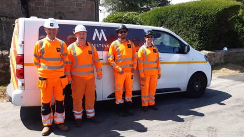 Four men wearing orange hi-vis outfits and white helmets are standing in front of a small white van with the word "multevo" on the front