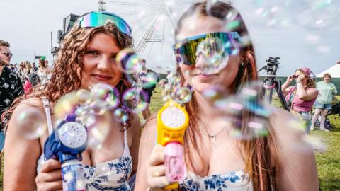 Two young women in matching floral blue and white strappy tops hold bubble guns, as bubbles shoot out around them. They are both wearing iridescent sunglasses, and are standing in a field with a ferris wheel behind them and other festivalgoers.