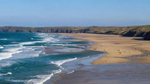 An aerial photo of Perranporth beach on a blue sky day. The waves are laced with white surf and patches of wet sand can be seen between sections of the waves. There are lifeguard cars parked on the beach. There are walkers along it and some people are swimming, surfing and paddling in the waves. The beach is lined with grass-topped sand dunes.