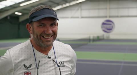 Neil Bamford is standing on the tennis courts and he is staring directly at the camera and laughing. He is wearing a black visor and white tennis T-shirt. 