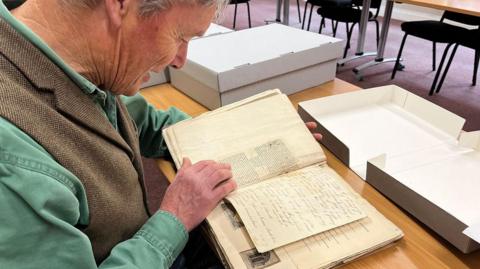 Tim Laycock holding an old manuscript book with ink handwriting and newspaper clippings inside