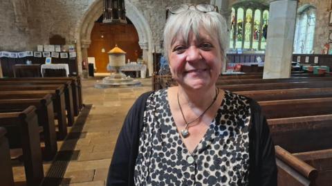 Deborah, wearing a blouse and black cardigan, smiles at the camera. She has grey hair and glasses on her head and is standing in an empty church.