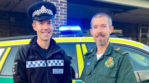 Dave Gardiner and paramedic Steve Gardiner stand together in uniform in front of an ambulance first responder car.