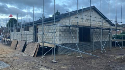 A picture of a building that is under construction. It has a grey brick shell and a roof, but there is still scaffolding around it. The sky in the background has grey clouds but also blue skies. In front of it the ground is muddy.