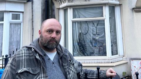 A concerned man stood outside his house with a sad look on his face, due to the damage on the property behind him. He has a bald head, beard and behind him is a cream terraced house.